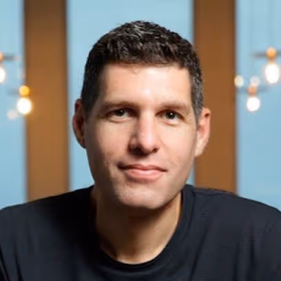 Portrait of a man with short dark hair wearing a black shirt, with blurred lights and blue panels in the background.