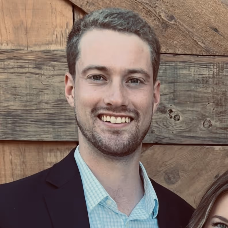 Smiling man with short brown hair and trimmed beard wearing a light blue checkered shirt and dark blazer standing in front of a wooden wall.