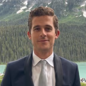 Man in a dark suit and light gray tie standing outdoors with a forest and lake in the background.